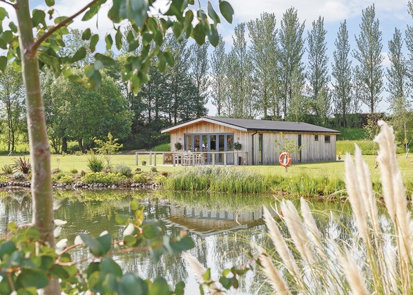 A wooden cabin with a deck, surrounded by a pond and tall trees.