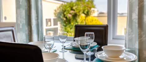 A dining table set with plates, bowls, wine glasses, and cutlery, with a view of greenery and buildings through the window.