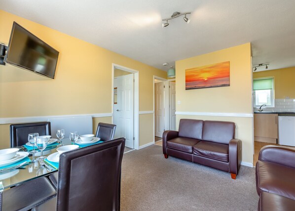 A dining area with a table set for four, a brown leather sofa, a flat-screen TV mounted on the wall, and a kitchen area with a window. - Gold 2 Bedroom Chalet Pet (Brixham, Torbay)