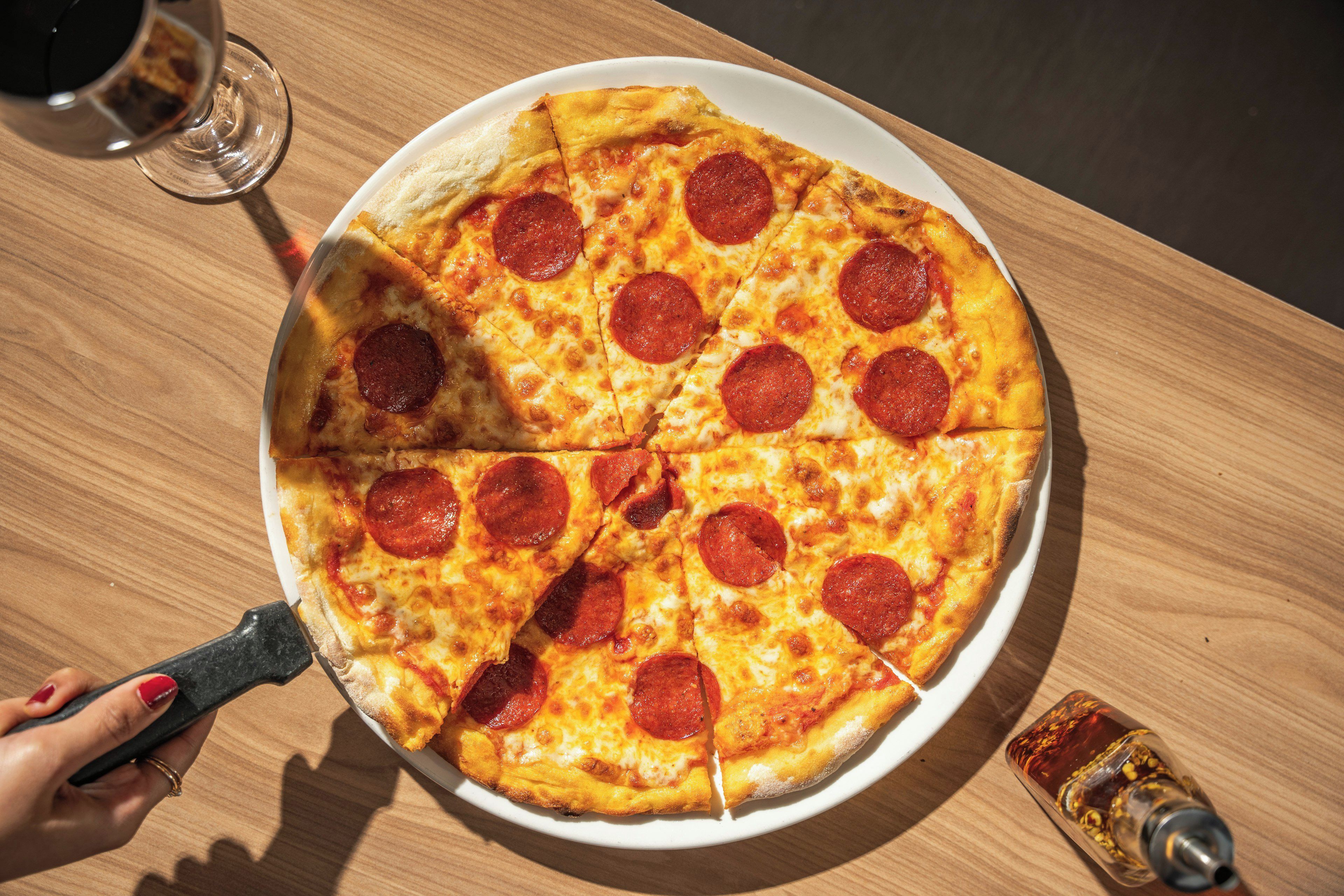 Una pizza de pepperoni sobre un plato blanco, con un cortador de pizza y una botella, todo sobre una mesa de madera.
