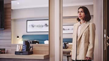 A woman in a beige blazer standing in a hotel room with a wooden desk, a coffee machine, and a framed picture on the wall.