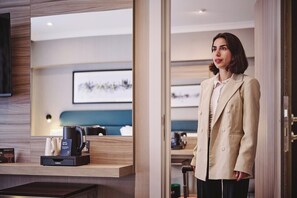 Una mujer con un blazer color beige parada en una habitación de hotel con un escritorio de madera, una cafetera y un cuadro en la pared.