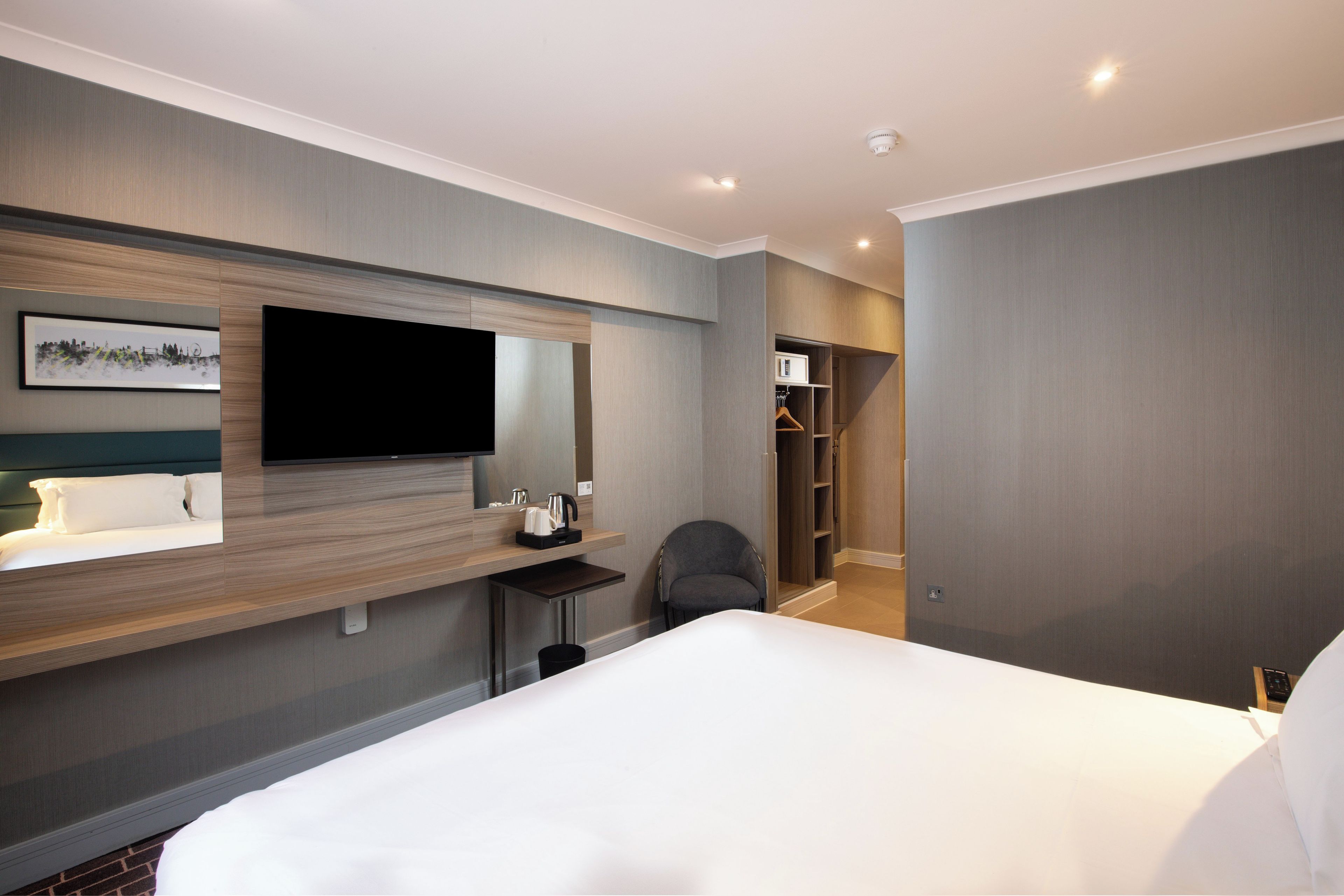 A modern hotel room with a large bed, a flat-screen TV mounted on the wall, a bedside table with a kettle, a chair, and a walk-in closet.