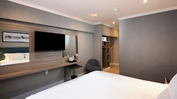 A modern hotel room with a large bed, a flat-screen TV mounted on the wall, a bedside table with a kettle, a chair, and a walk-in closet.