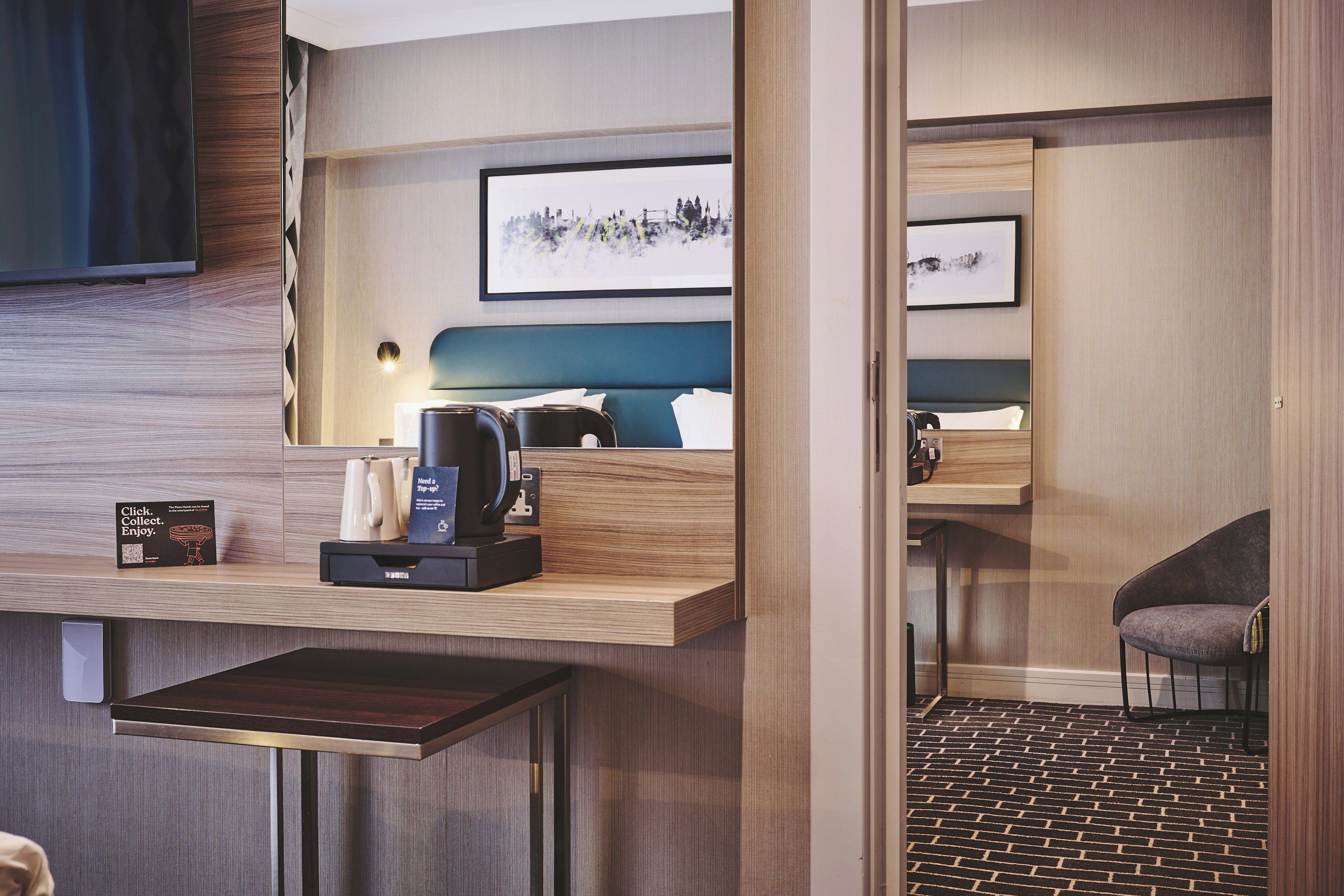 A modern hotel room with a wooden wall, a flat-screen TV, a bed with a blue headboard, a coffee machine, and a small table.