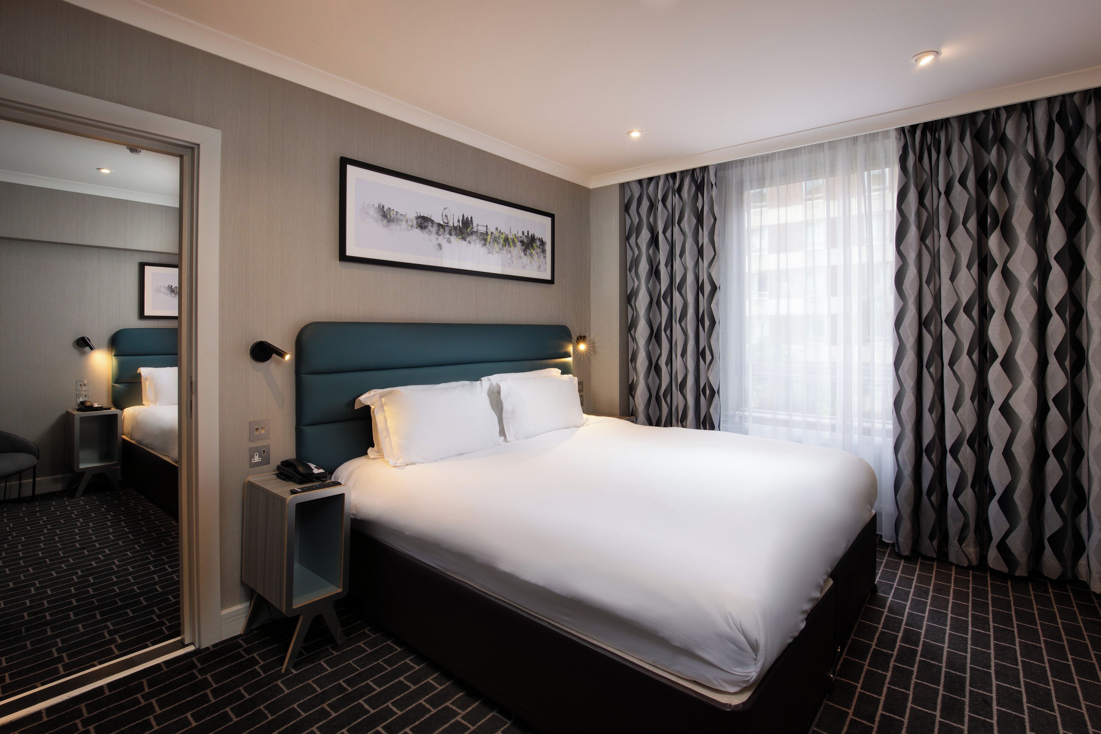 A hotel room with a large bed, a bedside table with a phone and remote, a chair, a mirror, and a window with patterned curtains.