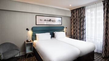 A hotel room with a double bed, a bedside table with a phone, a chair, a framed picture on the wall, and a patterned curtain.