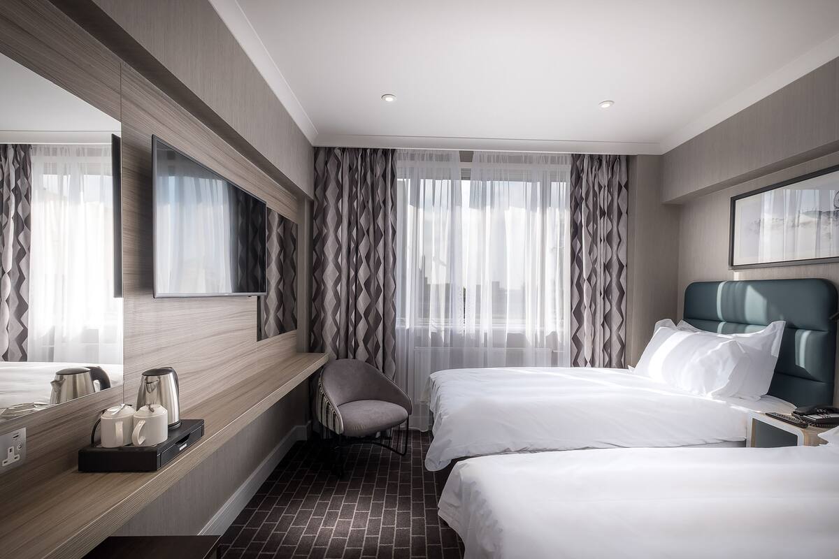 Ein Hotelzimmer mit zwei Betten, einem Schreibtisch mit Stuhl, einem großen Spiegel und einem Fenster mit leichtem Vorhang.