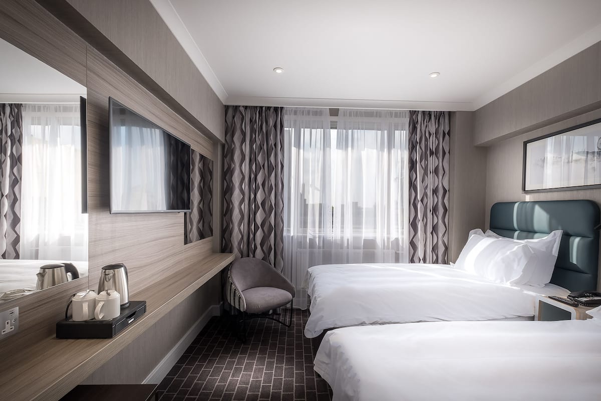 Une chambre d’hôtel avec deux lits, un bureau avec une chaise, un grand miroir et une fenêtre avec des rideaux légers.