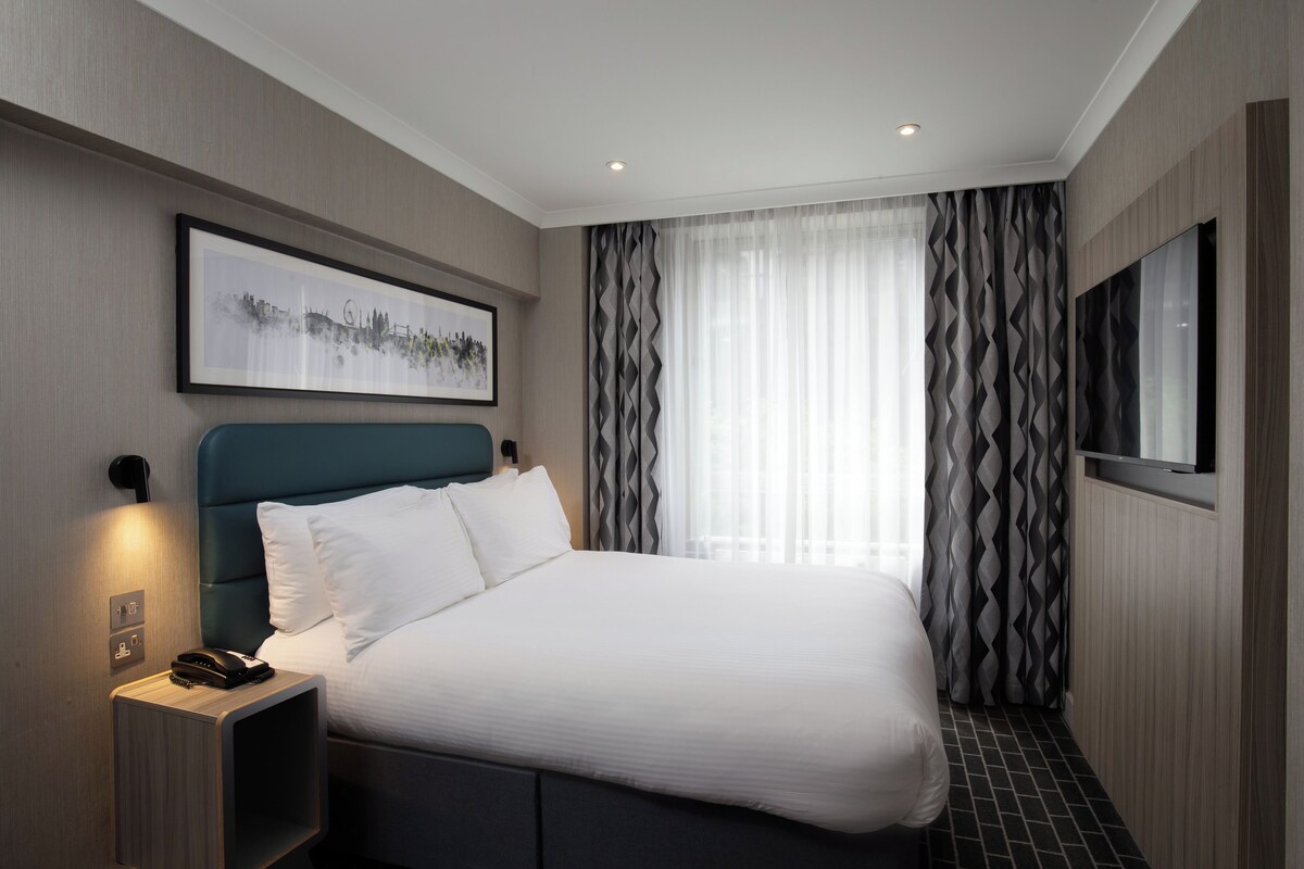 A modern hotel room with a large bed, a bedside table with a phone, a framed picture on the wall, and a TV on the wall.