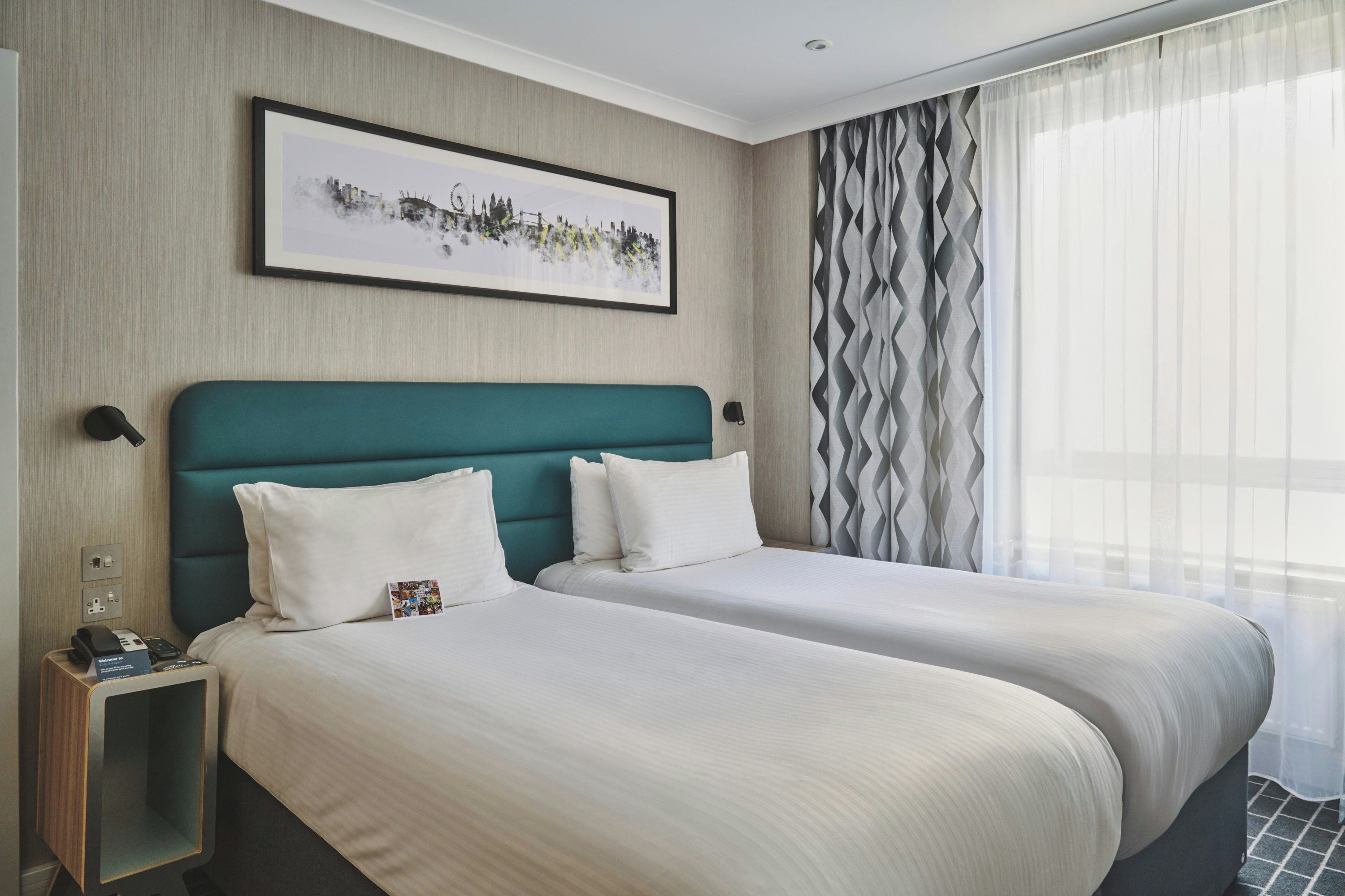 Ein Hotelzimmer mit zwei Betten, einem gerahmten Bild an der Wand, einem Nachttisch mit Telefon und Lampe sowie einem Fenster mit leichtem Vorhang.