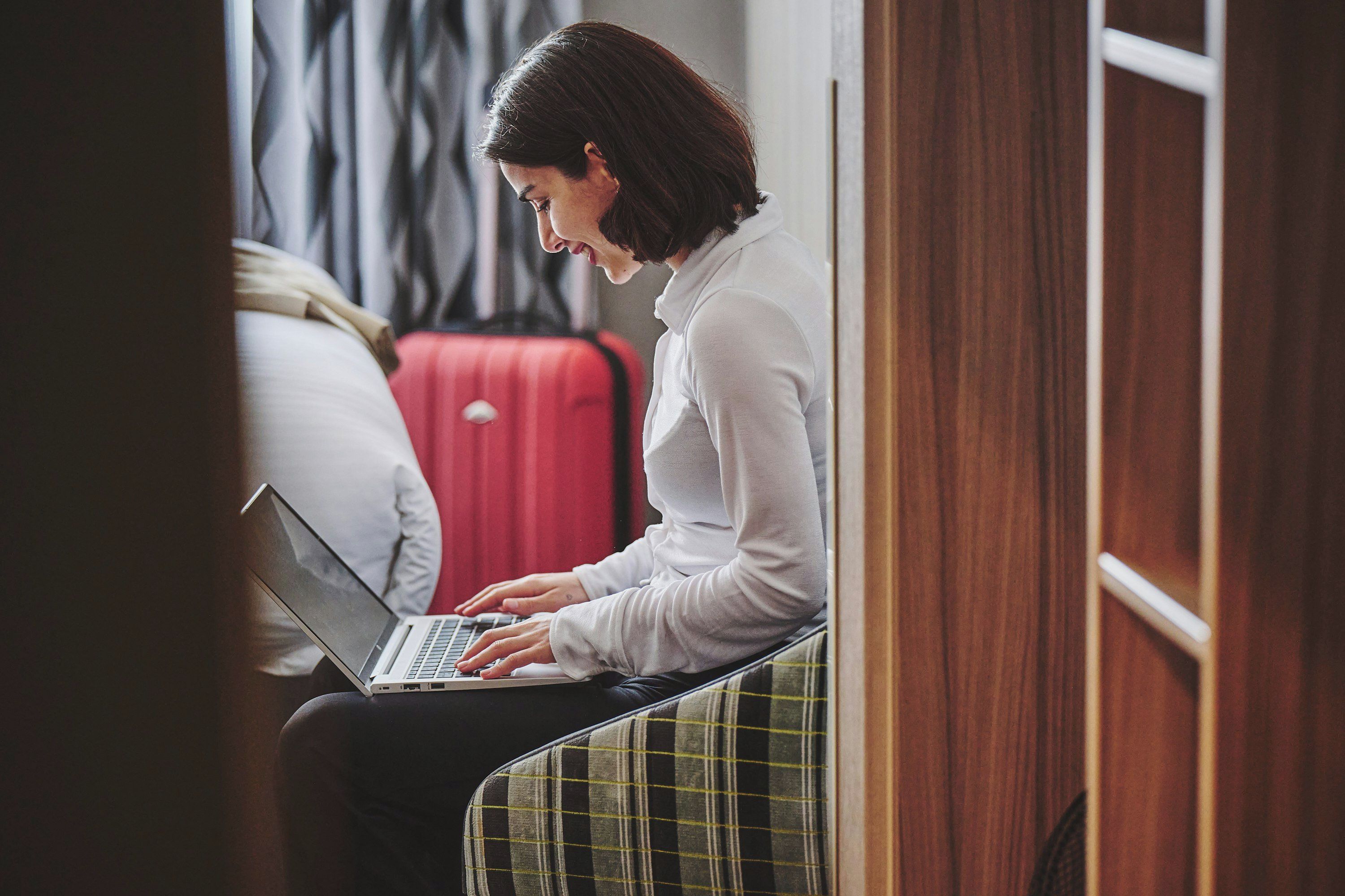 A person sitting on a bench with a laptop, a red suitcase, and a bed with a white pillow in the background.