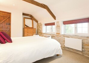 A bedroom with a large bed, wooden dresser, stone wall, window with red valance, and a wooden door.