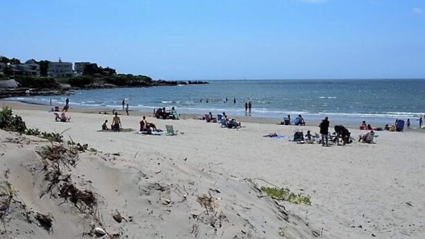 Une plage où l’on voit des gens se prélasser au soleil et nager, un rivage rocheux et des bâtiments en arrière-plan.