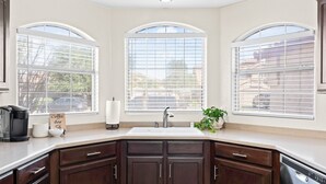 A kitchen with dark wooden cabinets, a white countertop, a sink, and three large windows with blinds. - Sunny Peoria Escape w/ Private Pool + Fire Pit (Peoria)