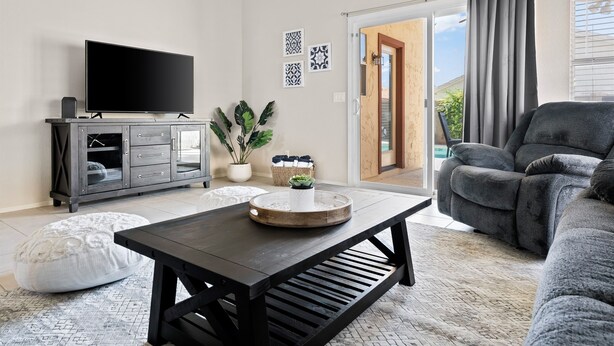 A modern living room with a flat-screen TV, dark wooden coffee table, grey sofa, and a sliding glass door leading to a balcony. - Sunny Peoria Escape w/ Private Pool + Fire Pit (Peoria)