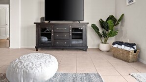 A living room with a dark wooden TV stand, a flat-screen TV, a white pouf, a potted plant, and a woven basket. - Sunny Peoria Escape w/ Private Pool + Fire Pit (Peoria)