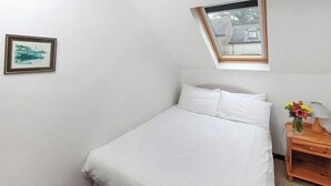 Un dormitorio con cama, una mesita de noche con lámpara, un cuadro enmarcado en la pared y un tragaluz.