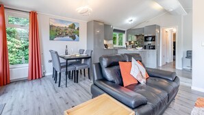A modern living room with a leather sofa, dining table, and chairs, a kitchen in the background, and a painting on the wall. - Bakewell Deluxe Lodge (Longnor, Nr. Buxton)