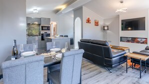 A modern living room with a dining area, a black leather sofa, a flat-screen TV, and a kitchen in the background. - Bakewell Deluxe Lodge (Longnor, Nr. Buxton)