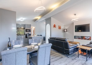 A modern living room with a dining area, a black leather sofa, a flat-screen TV, and a kitchen in the background.