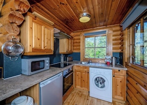 A compact kitchen with wooden cabinets, a microwave, a dishwasher, and a window with a view of greenery. - Timber lodge set in an elevated position with views of the lake. (Webbery, Bideford)