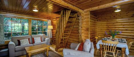 Interior de una cabaña de troncos con un sofá, sillones, una mesa de comedor y vistas al bosque a través de amplias ventanas.