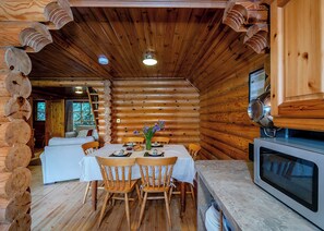 A log cabin interior with a dining area, wooden chairs, a table set for two, and a kitchen with a microwave. - Timber lodge set in an elevated position with views of the lake. (Webbery, Bideford)
