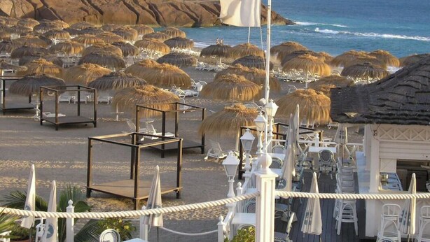 A beachside hotel with thatched umbrellas, lounge chairs, and a flag. - Costa Adeje Holiday Apartment (Adeje)