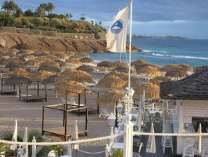 A beachside hotel with thatched umbrellas, lounge chairs, and a flag. - Costa Adeje Holiday Apartment (Adeje)