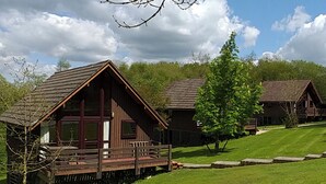 A row of wooden cabins with a deck, surrounded by a grassy area and trees. - Single storey A-frame lodge. (North Tamerton, Bude)