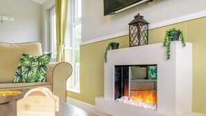 Una sala de estar con chimenea, un sofá con cojín estampado de motivos tropicales, una mesita de madera y un televisor montado en la pared.