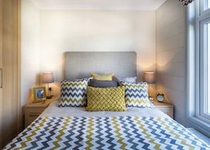 Uma cama bem arrumada com colcha estampada, várias almofadas, abajures e um quadro na mesinha de cabeceira.