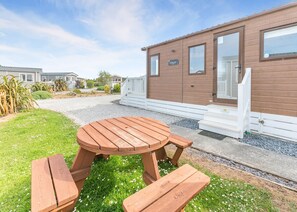 Eine Holzterrasse mit einem runden Tisch und Bänken, eine weiße Veranda und ein Gebäude mit Fenstern und einer Tür.