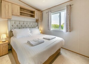 A neatly made bed with white linens, a bedside lamp, and a window with curtains. - Luxurious 14ft-wide holiday home. (Looe)