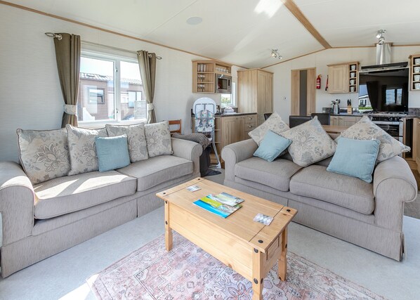 A living room with two sofas, a coffee table, and a kitchen area with cabinets and appliances. - Luxurious 14ft-wide holiday home. (Looe)