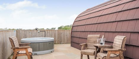 A hot tub, wooden chairs, and a table with glasses and a bottle on a patio with stone tiles.