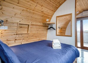 A room with a wooden ceiling, a bed with a blue mattress, a mirror, and a window.