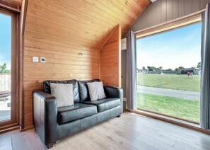 A room with a wooden ceiling, a black leather sofa with cushions, a large window, and a view of a grassy area outside.