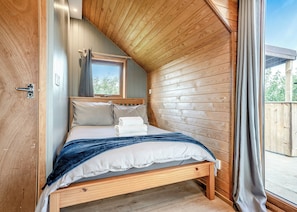 A bedroom with a wooden ceiling, a window, a bed with pillows and a folded towel, and a wooden door.