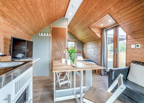 A modern cabin interior with a wooden ceiling, a dining area with a table and chairs, a seating area with a sofa, a television, and a window with a view of the outdoors.