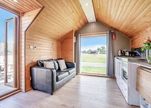 A cozy room with a wooden ceiling, a black leather sofa, a kitchen area with modern appliances, and a large window offering a view of the outdoors.