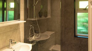 Une salle de bain équipée d’un lavabo, d’un miroir, d’une douche et d’une fenêtre donnant sur un espace verdoyant.