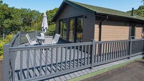 Une terrasse extérieure couverte, avec une table et des chaises, un bâtiment en bois doté d’un toit végétalisé et une rambarde en métal.