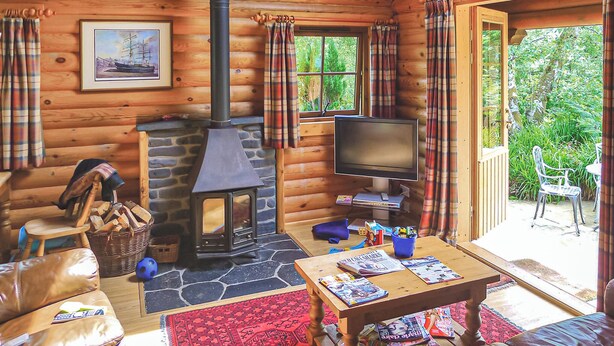 A log cabin interior with a fireplace, television, seating area, and a view of greenery outside.