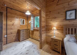 Ein Zimmer in einer Holzhütte mit einem Bett, einem Nachttisch, einem Schminktisch, einem Spiegel, einem Fenster mit Blick auf Bäume und einer Deckenleuchte.