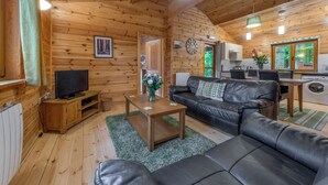 A log cabin interior with a living room, a television, a sofa, a coffee table, a kitchen area, and a window with a view of trees. - 2 bedroom accommodation in Ellesmere (Ellesmere)