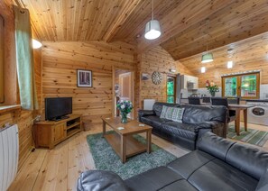 A log cabin interior with a living room, a television, a sofa, a coffee table, a kitchen area, and a window with a view of trees. - 2 bedroom accommodation in Ellesmere (Ellesmere)