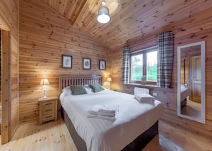 A wooden-paneled bedroom with a bed, bedside lamps, a window with curtains, and framed pictures on the wall.