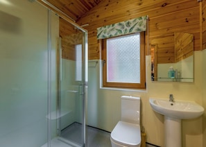 A bathroom with a glass shower enclosure, a toilet, a sink, and a window with a leaf-patterned curtain.